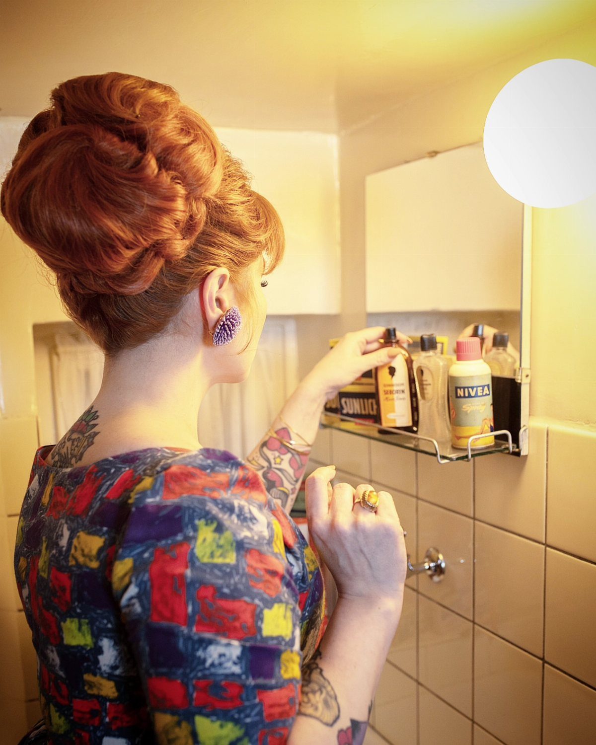1960s Hairstyles: 7 Ideas from Beehives to Bubble Cuts 2 Woman with a red beehive hairstyle reaching for a bottle in a bathroom.
