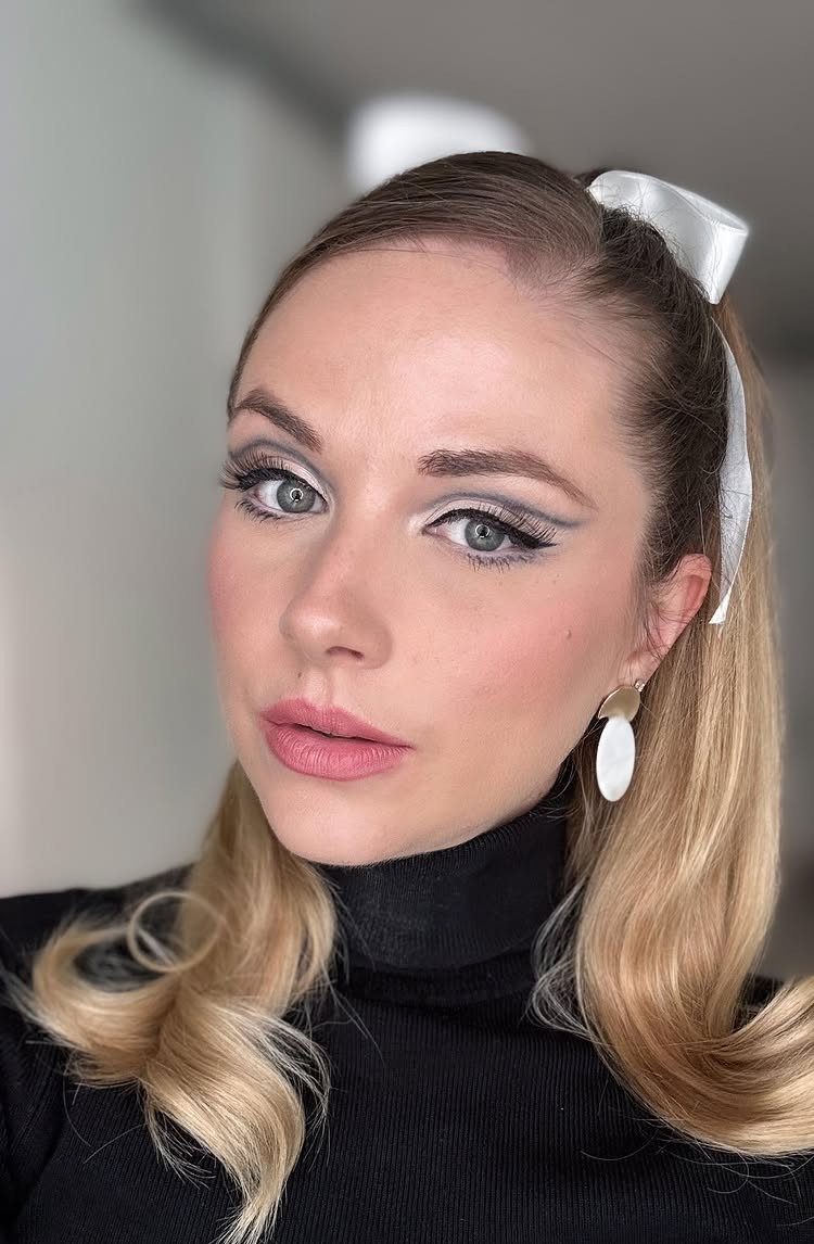 1960s Hairstyles: 7 Ideas from Beehives to Bubble Cuts 6 Woman with blonde hair wearing a white satin bow hair accessory and winged eyeliner.