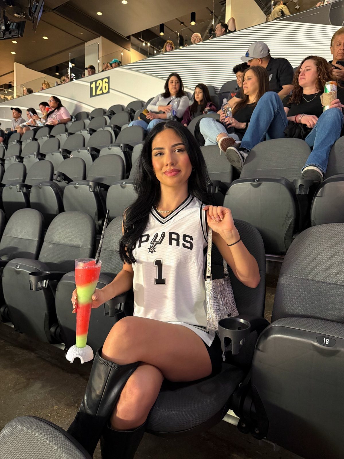Woman in a white Spurs basketball jersey and black boots sitting in stadium seats.