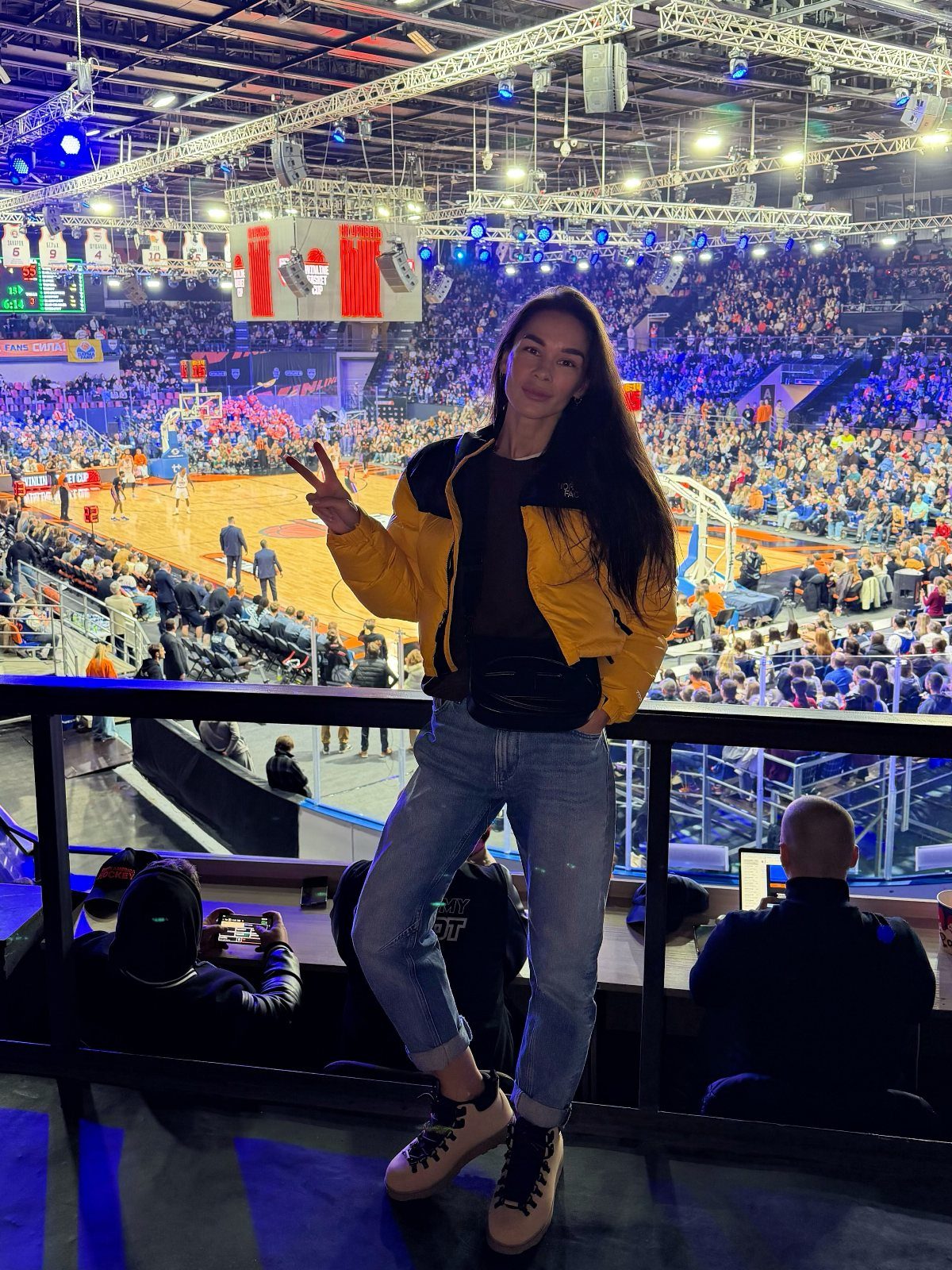 Woman in yellow puffer jacket and jeans posing at a basketball game arena