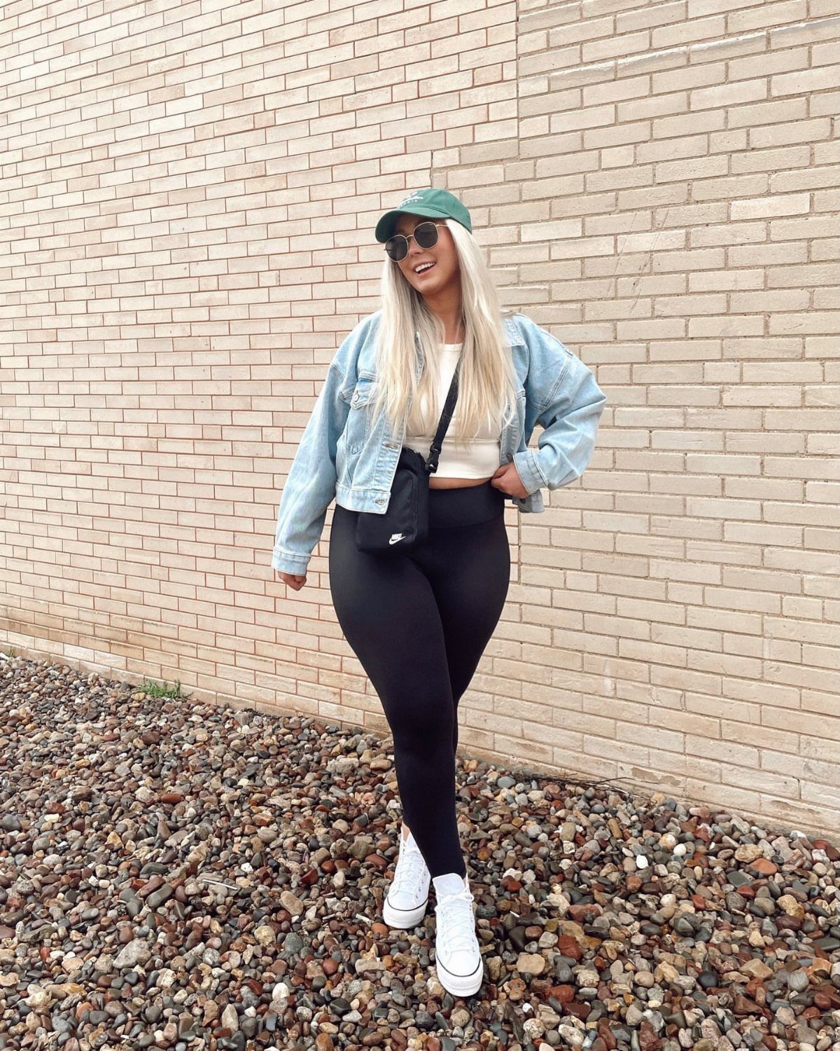 Woman wearing a denim jacket, black leggings, and white sneakers against a brick wall.