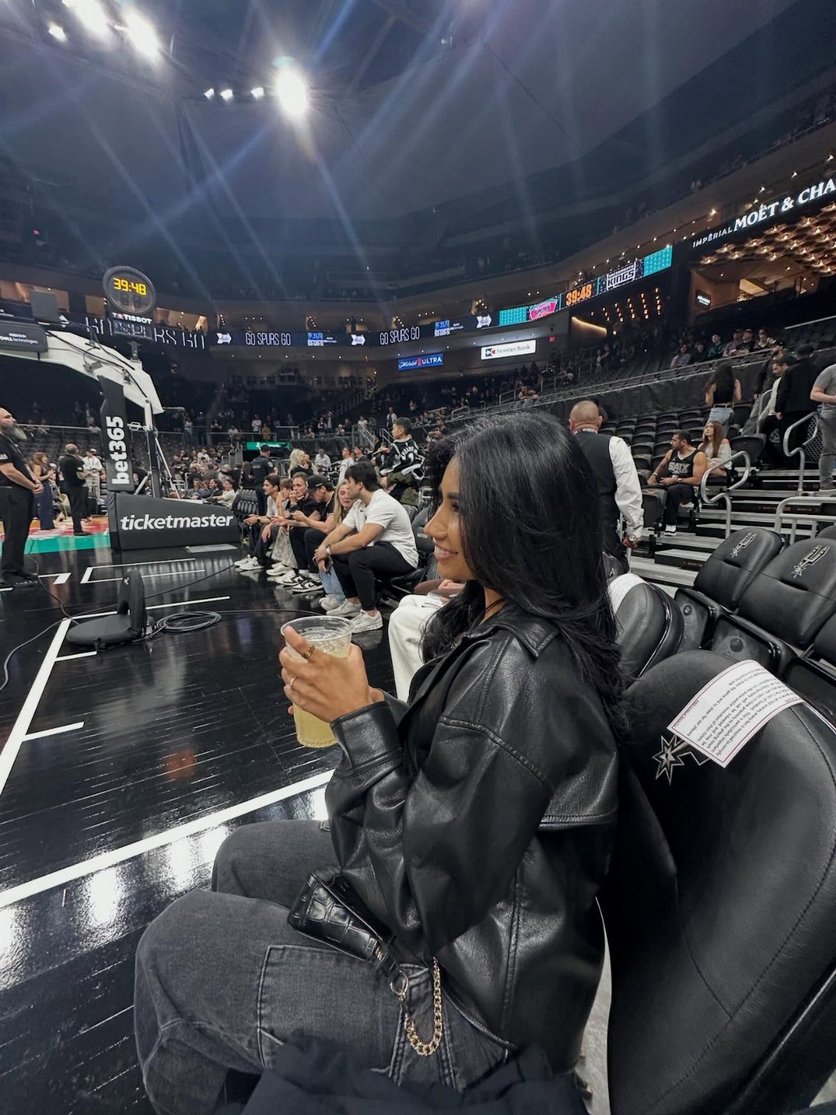 Woman in a black leather jacket and grey jeans sitting at a basketball game.