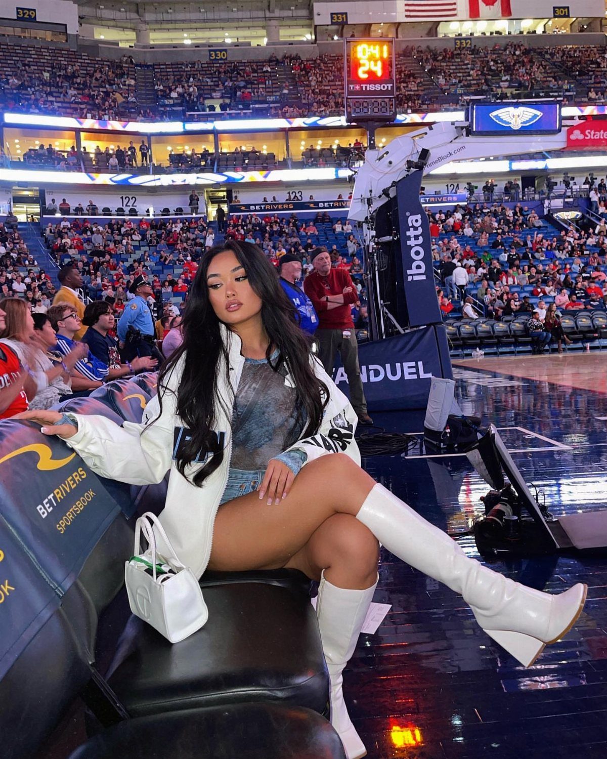 Woman sitting courtside wearing a denim skirt and white heeled knee-high boots.
