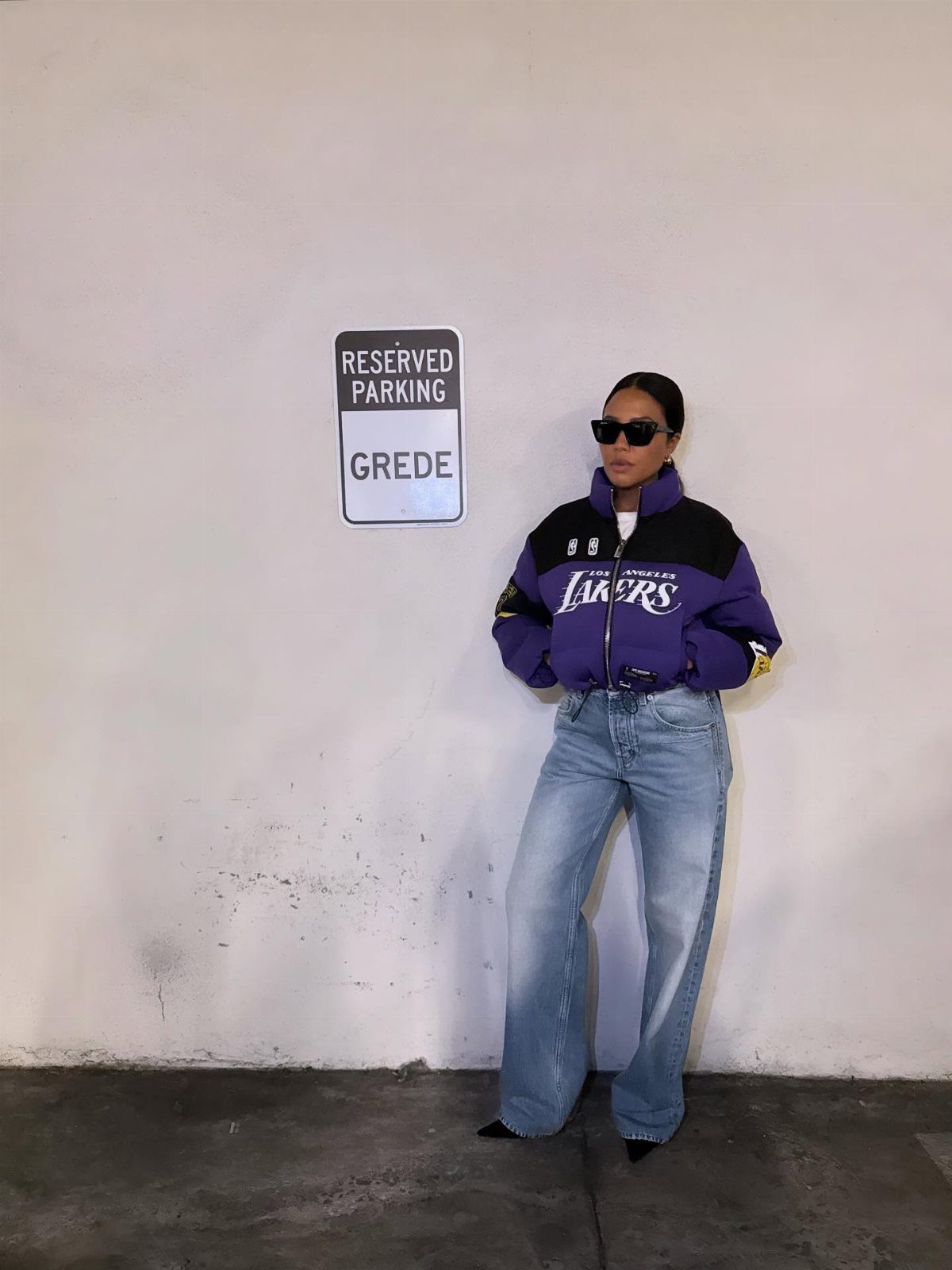 Woman in purple Lakers jacket and wide-leg jeans posing by a white wall