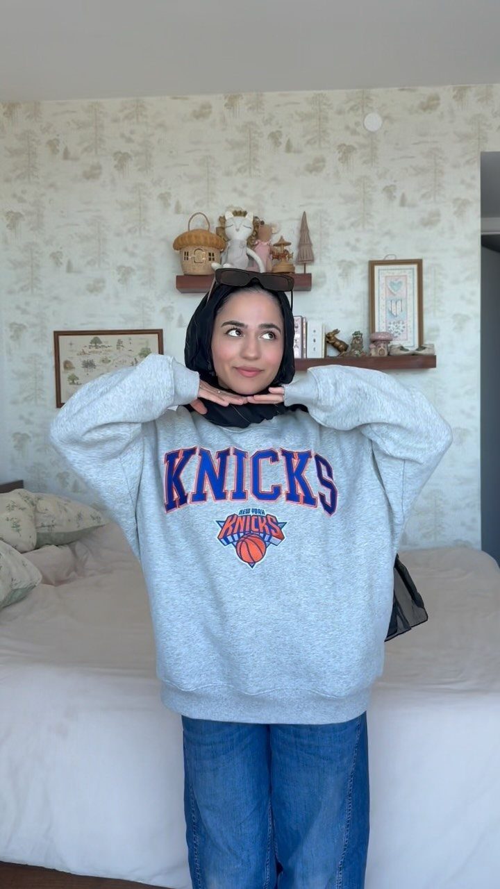 Woman wearing a grey New York Knicks sweatshirt, blue jeans, and a black hijab.