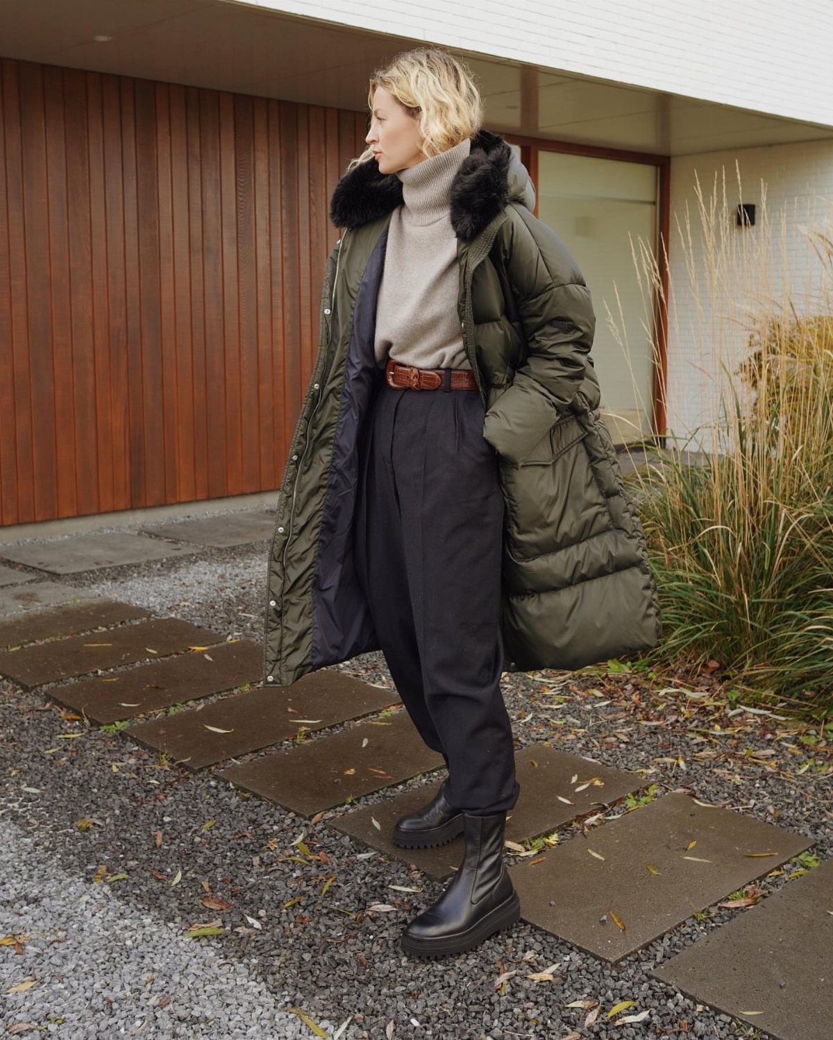 Woman in olive puffer coat and black baggy pants standing on stone path.