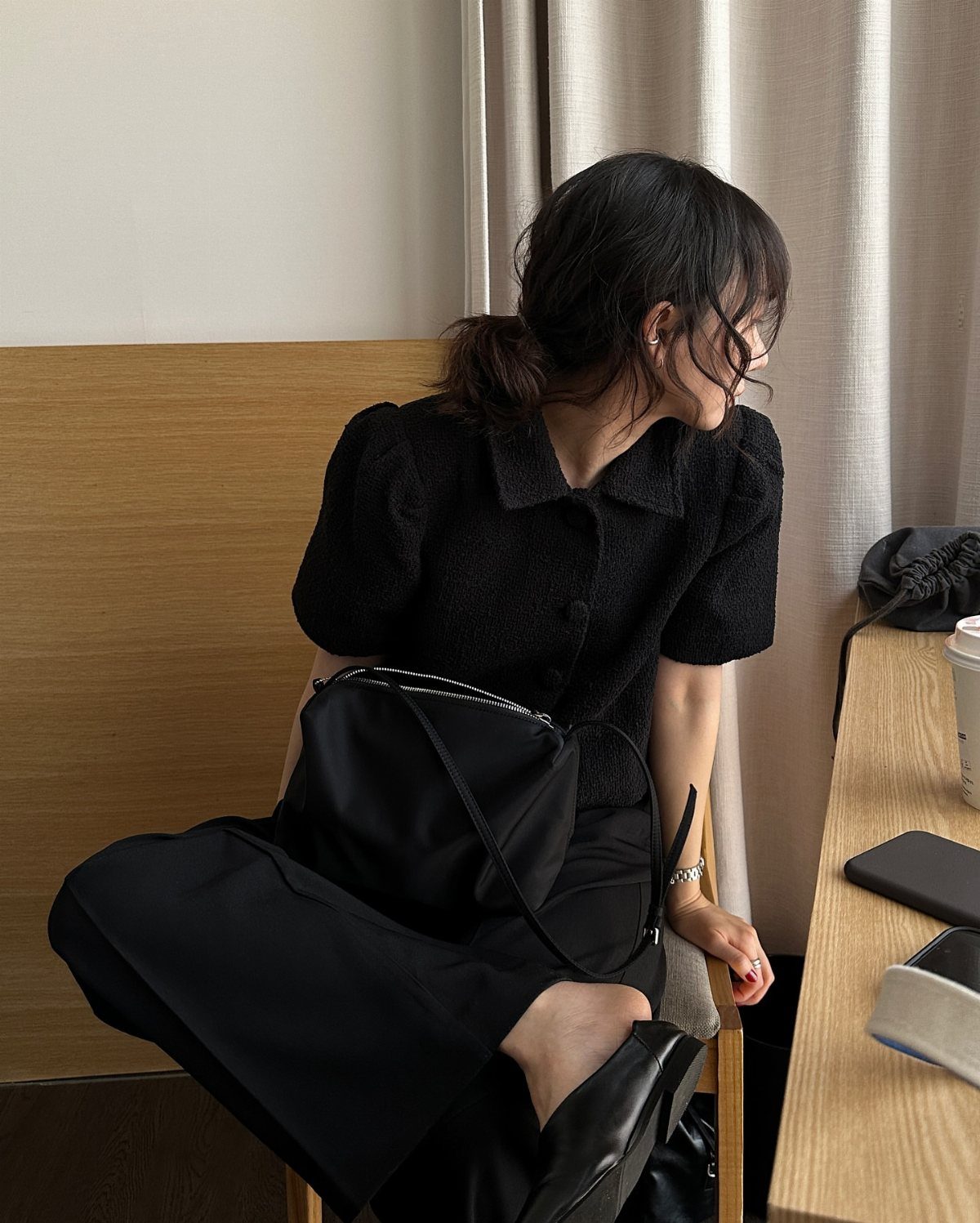Woman wearing a black buttoned tweed top and baggy black trousers sitting indoors.