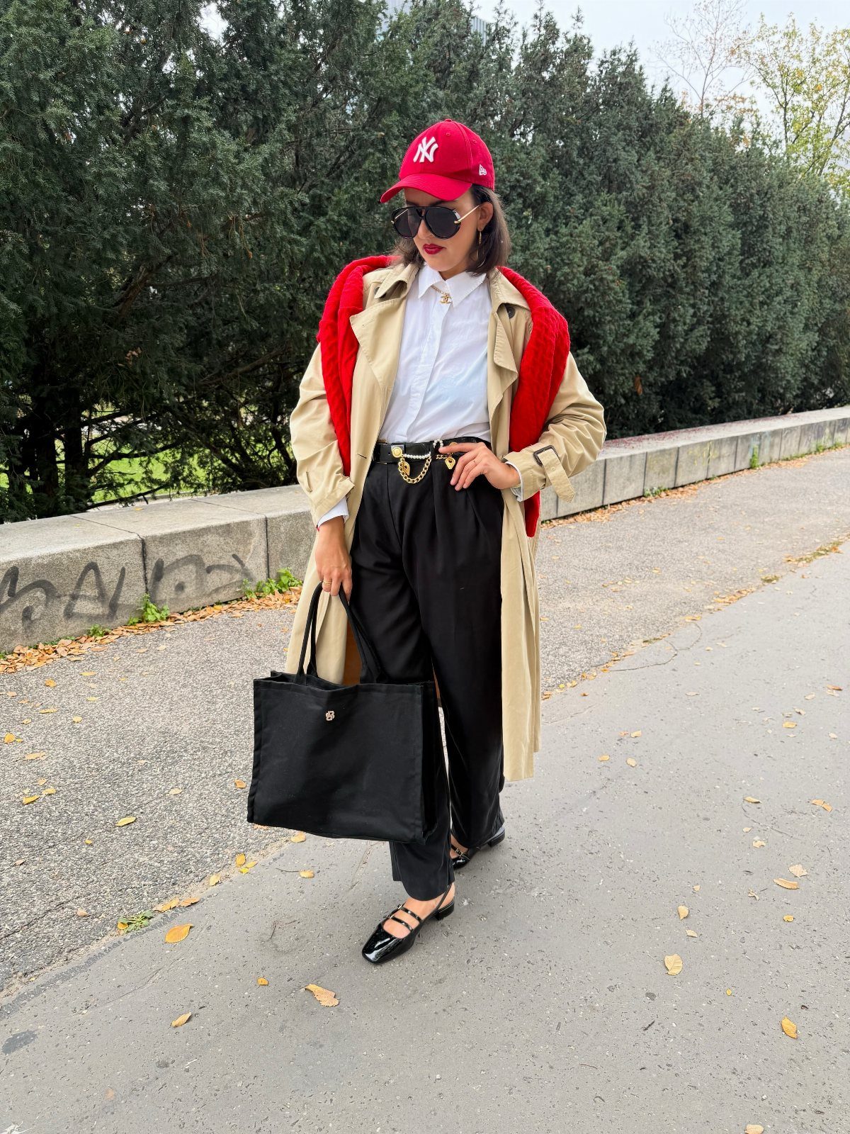 Woman in beige trench coat, white shirt, black baggy pants, and red accessories.