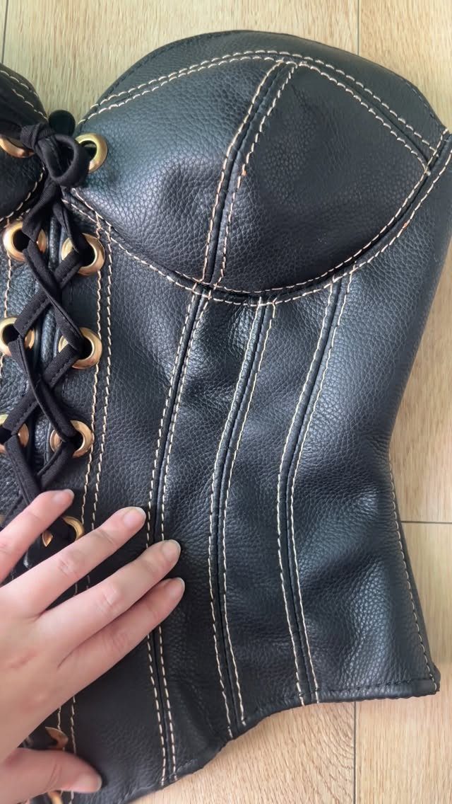 Black leather corset with white stitching and gold eyelets on a wooden floor
