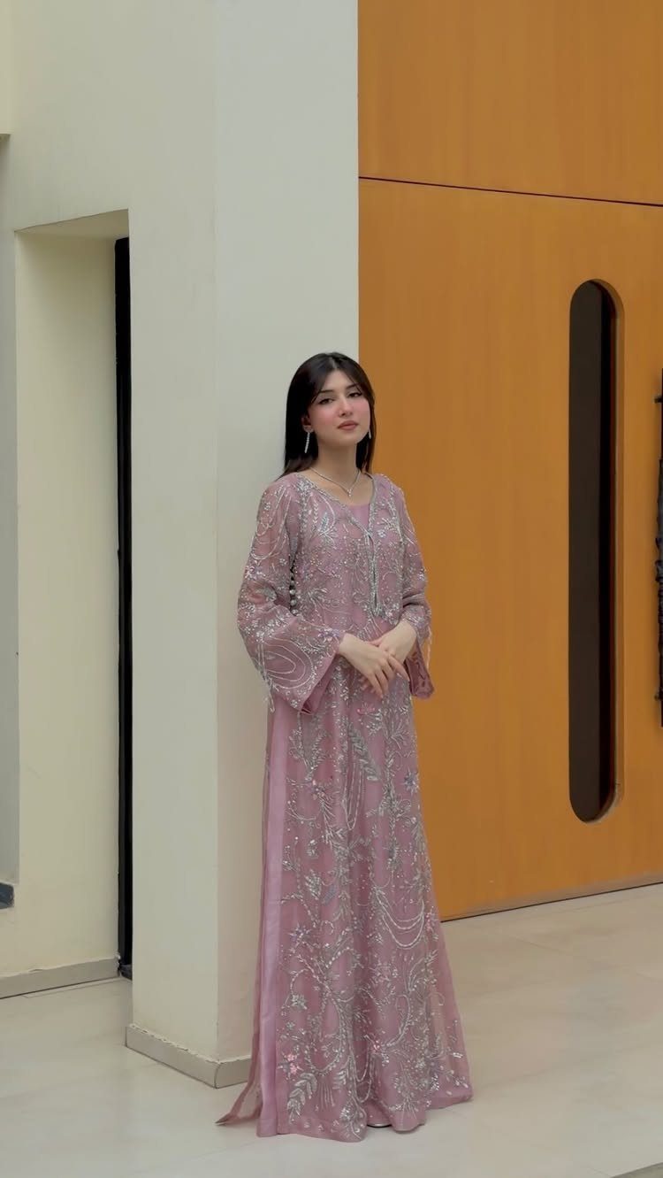 25 Blush Wedding Dress Ideas in Lace, Tulle, Silk and Sequins 19 Woman in a long-sleeved blush pink embroidered gown standing in a modern hallway.