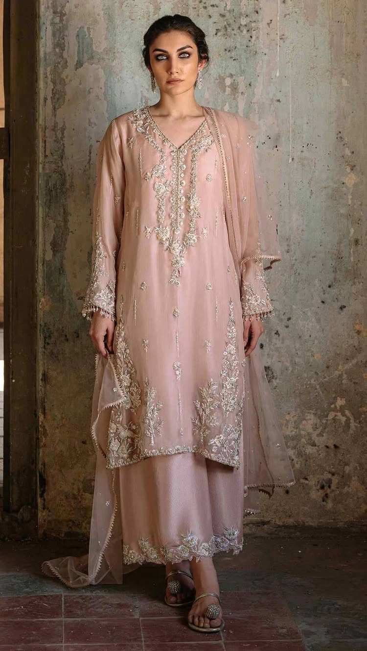 25 Blush Wedding Dress Ideas in Lace, Tulle, Silk and Sequins 26 Woman in a blush-colored embroidered traditional bridal outfit standing against a grey wall.