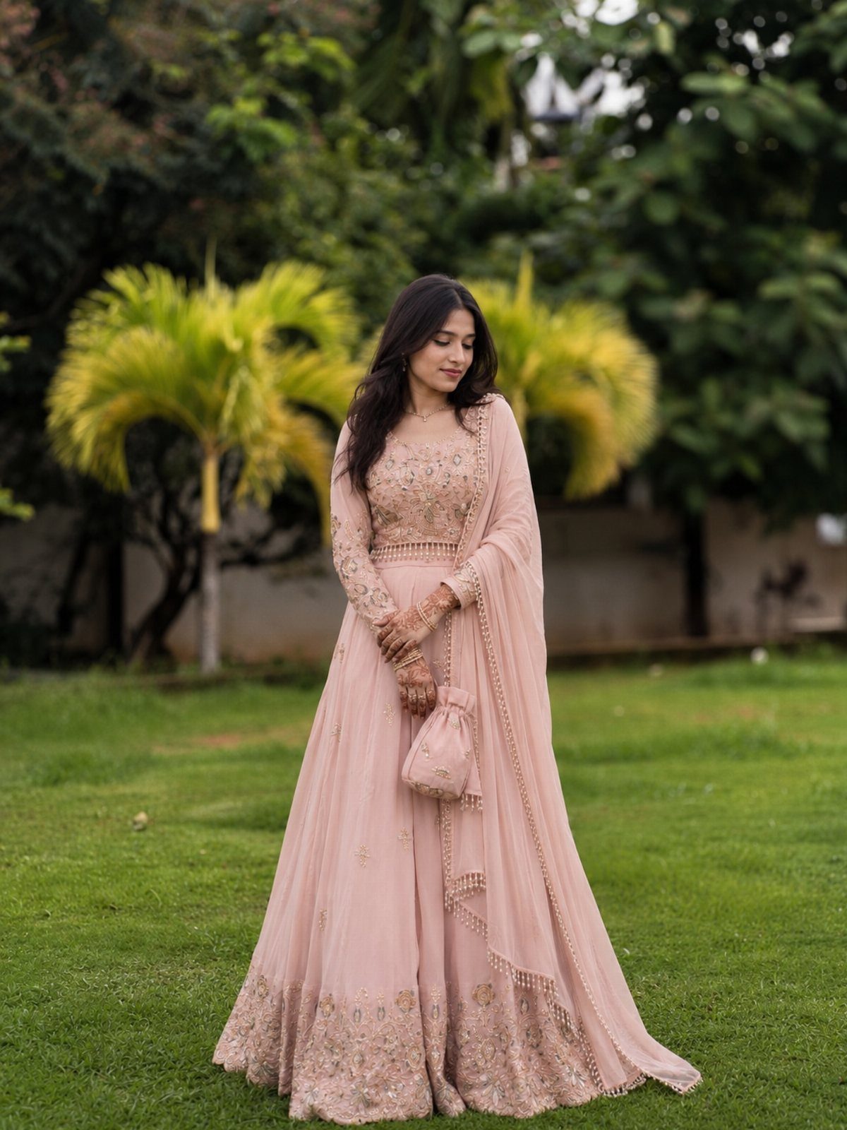 25 Blush Wedding Dress Ideas in Lace, Tulle, Silk and Sequins 14 Woman in a blush pink embroidered lehenga and dupatta standing on a green lawn.