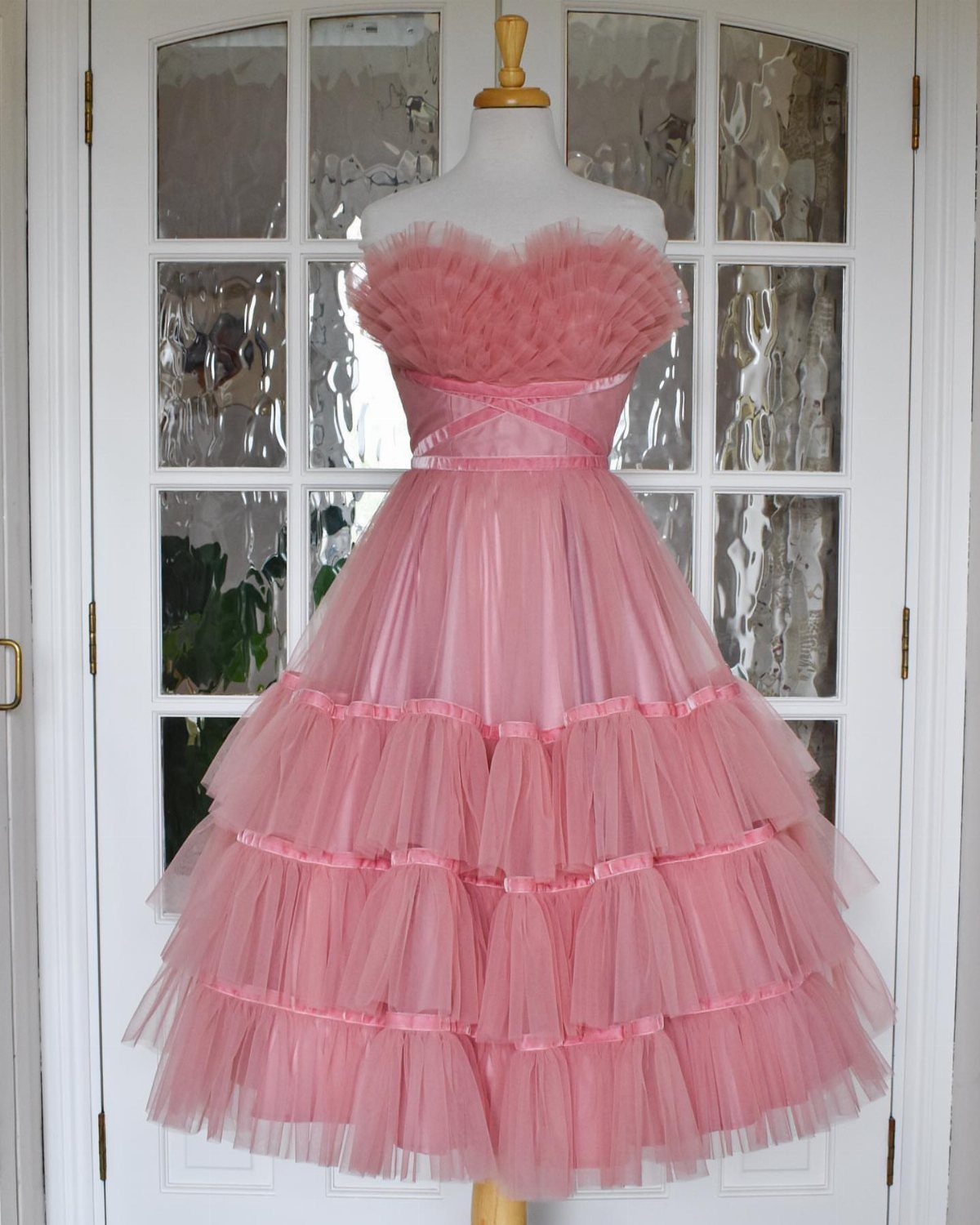 25 Blush Wedding Dress Ideas in Lace, Tulle, Silk and Sequins 11 Pink tiered tulle dress with ruffles on a white mannequin against white doors