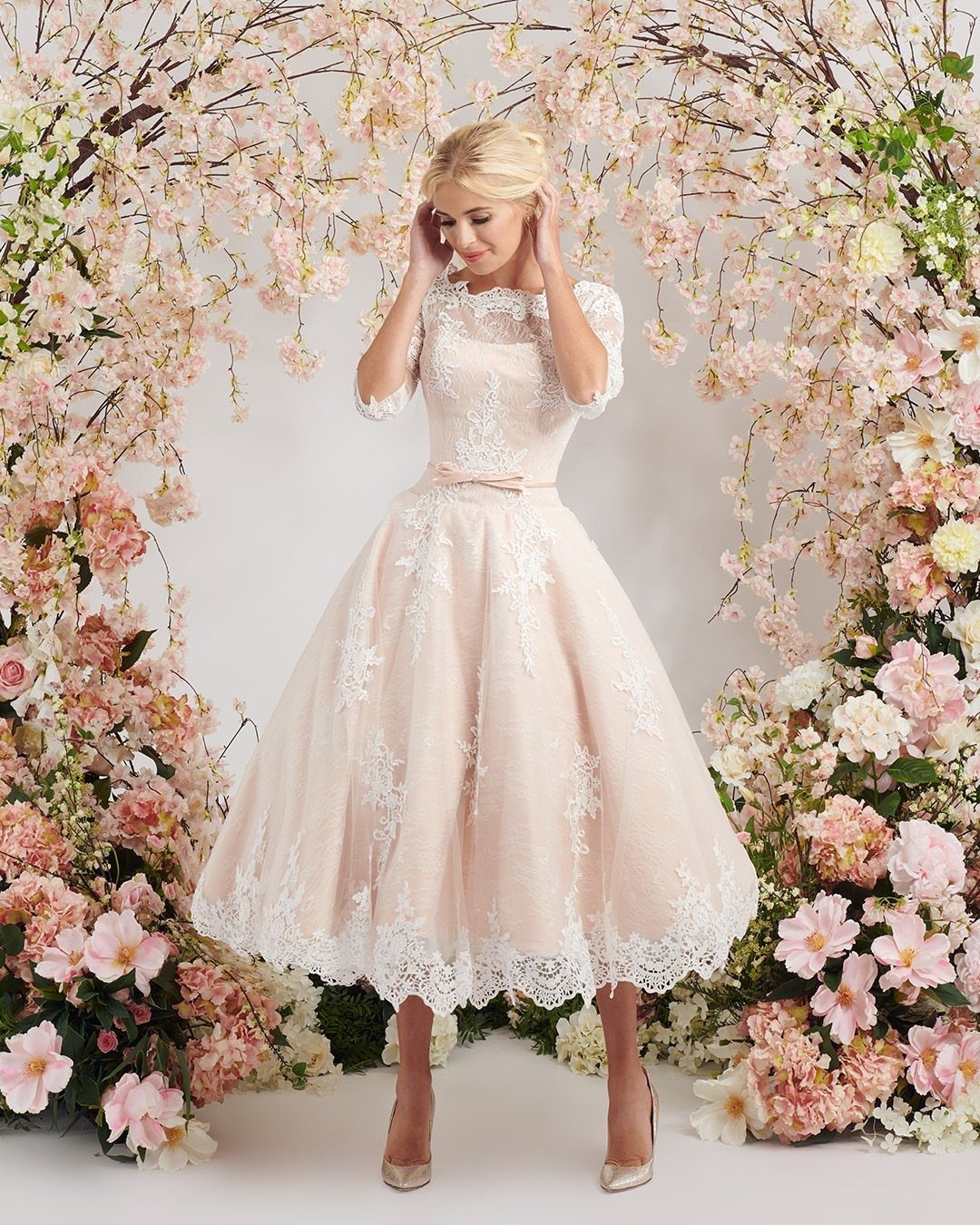 25 Blush Wedding Dress Ideas in Lace, Tulle, Silk and Sequins 22 Blonde woman in a blush lace wedding dress standing before a floral arch