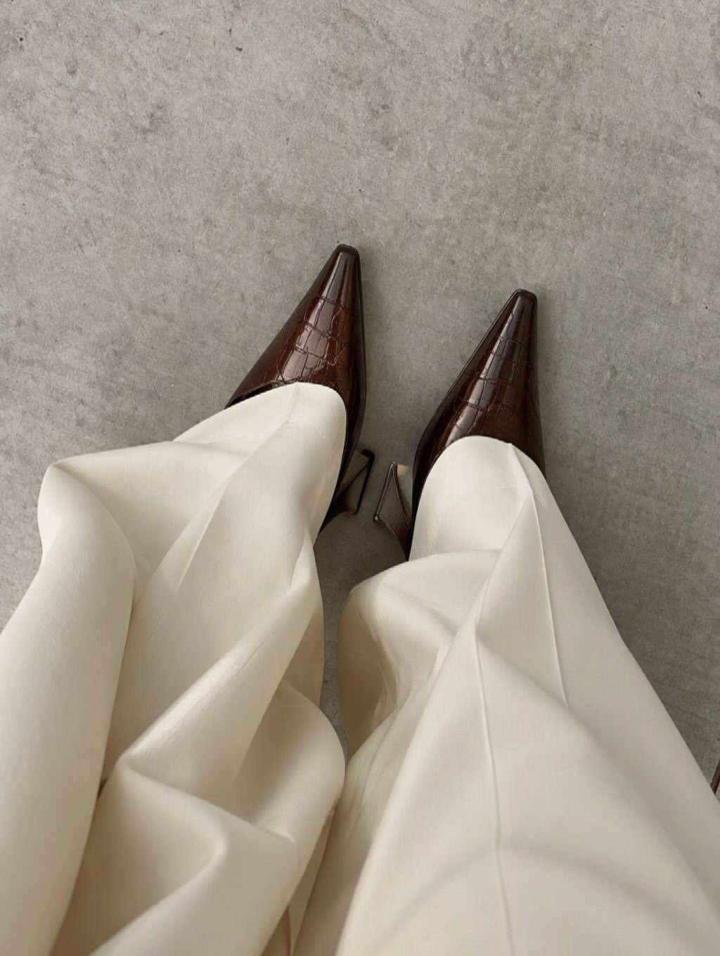 Brown pointed-toe slingback heels paired with cream wide-leg trousers on a grey floor.