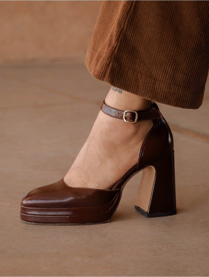 Brown glossy leather platform pumps with block heels and ankle straps worn with trousers.