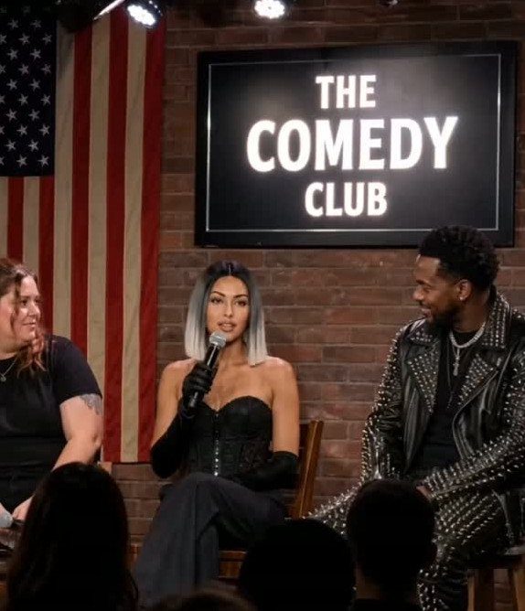 Woman in black corset and trousers sitting in a comedy club setting.