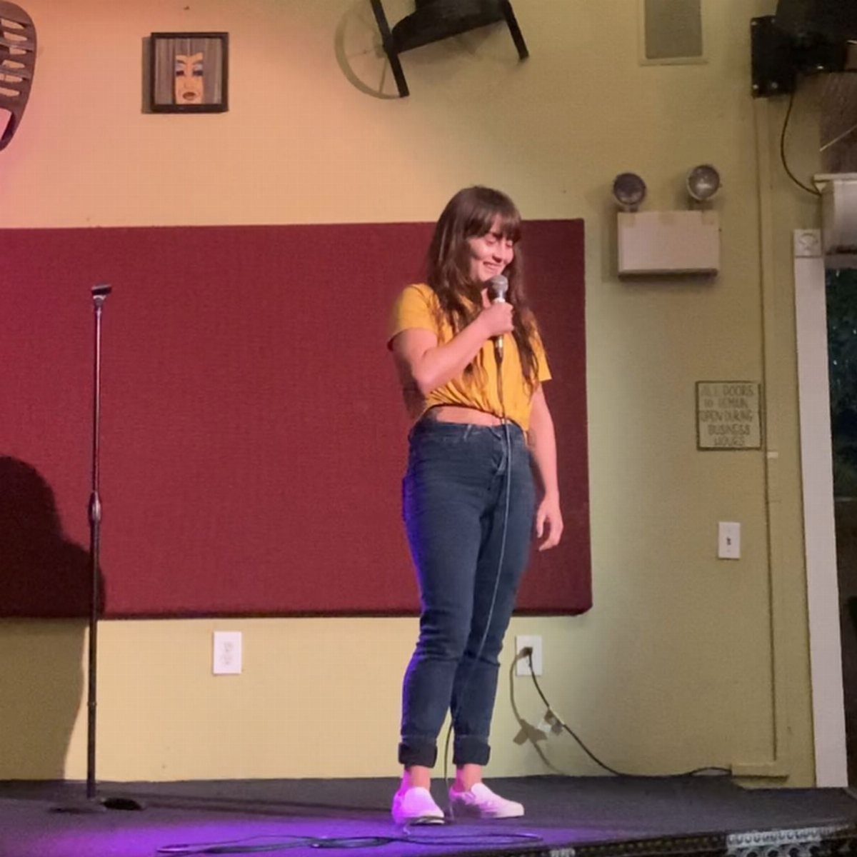 Woman on a comedy stage wearing a yellow crop top and high-waisted jeans.