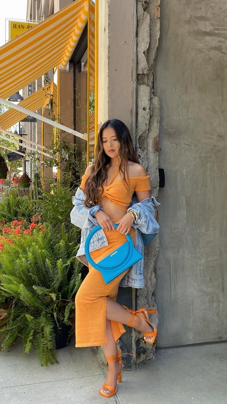 Woman in orange midi skirt and crop top with denim jacket and blue handbag.