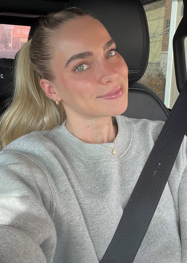 Woman with blonde hair in a high ponytail wearing a grey sweatshirt in a car.