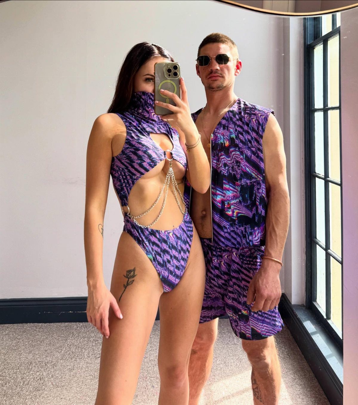 Man and woman in matching purple psychedelic print rave outfits posing for a mirror selfie.