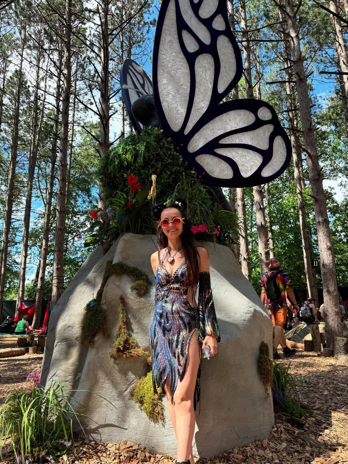 Woman in iridescent rave dress posing before a giant butterfly sculpture in woods
