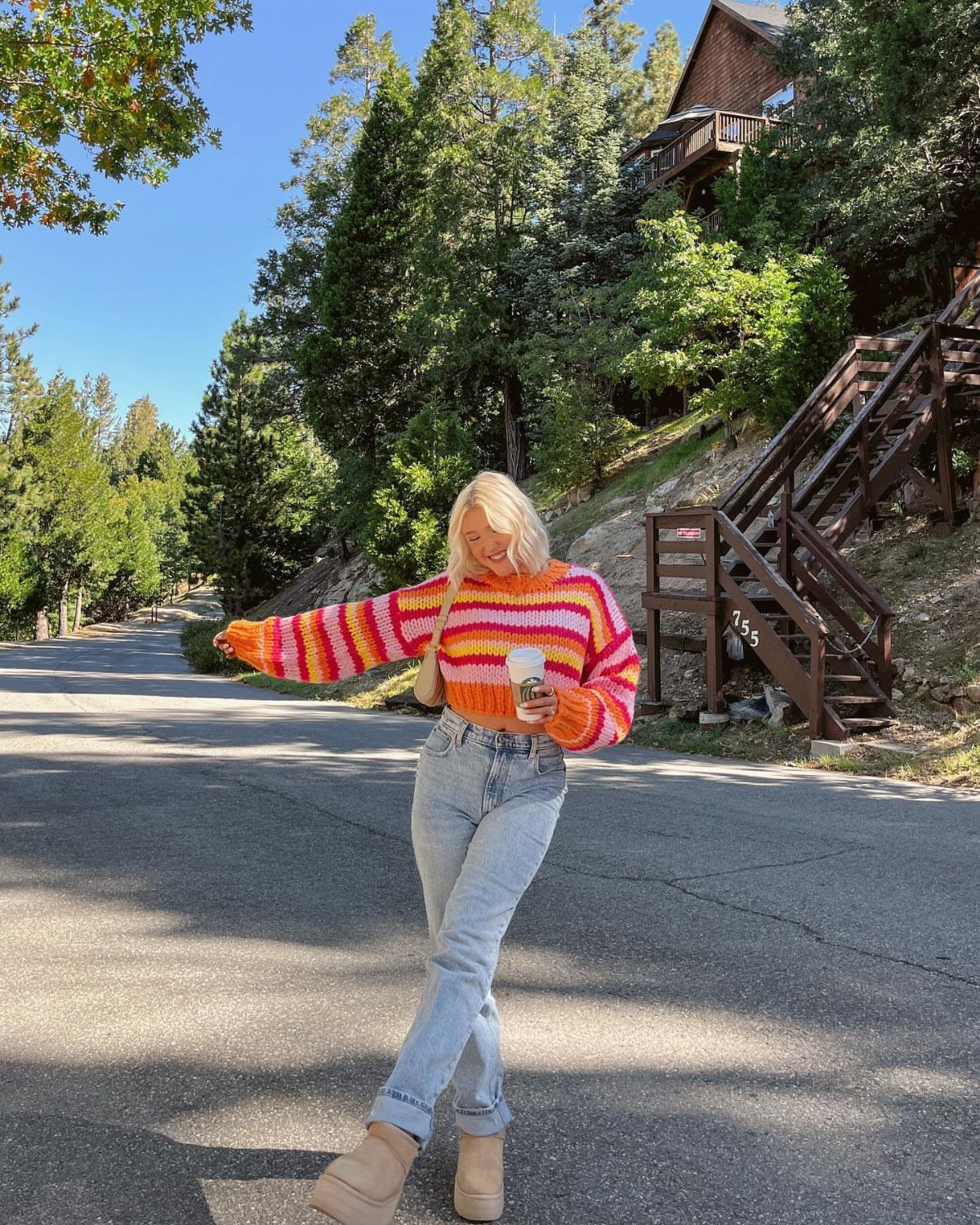 29 Fall Outfits Teens Can Copy, From Grunge to Cowgirl Looks 23 Woman in a bright striped sweater and blue jeans holding coffee on a road.