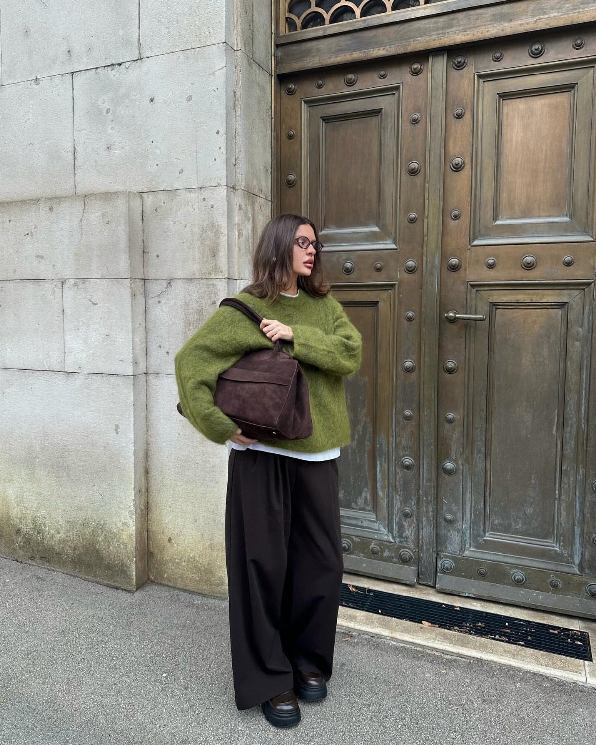 29 Fall Outfits Teens Can Copy, From Grunge to Cowgirl Looks 20 Woman wearing an olive green fuzzy sweater and wide black trousers by metal doors