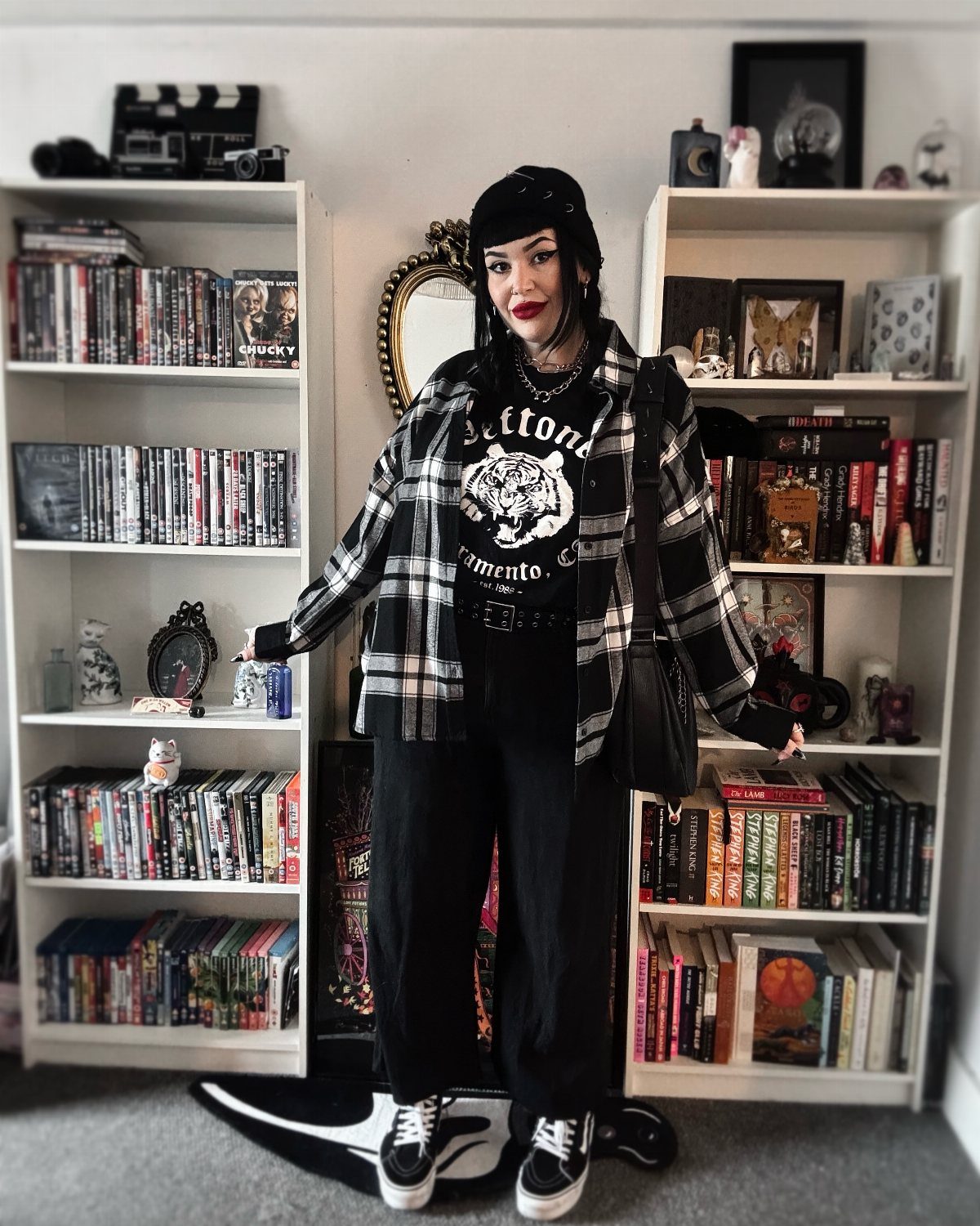 29 Fall Outfits Teens Can Copy, From Grunge to Cowgirl Looks 25 Young woman wearing a plaid shirt, graphic tee, and black trousers in a room.