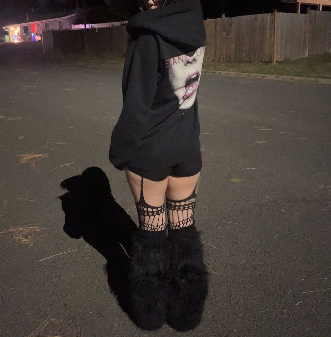 29 Fall Outfits Teens Can Copy, From Grunge to Cowgirl Looks 12 Person in oversized black graphic hoodie, fishnet stockings, and fluffy black boots at night.