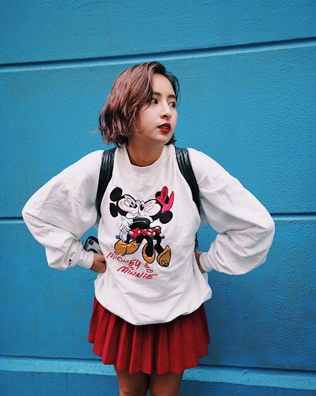 29 Fall Outfits Teens Can Copy, From Grunge to Cowgirl Looks 15 Young woman in a Mickey Mouse t-shirt and red pleated skirt against blue wall.