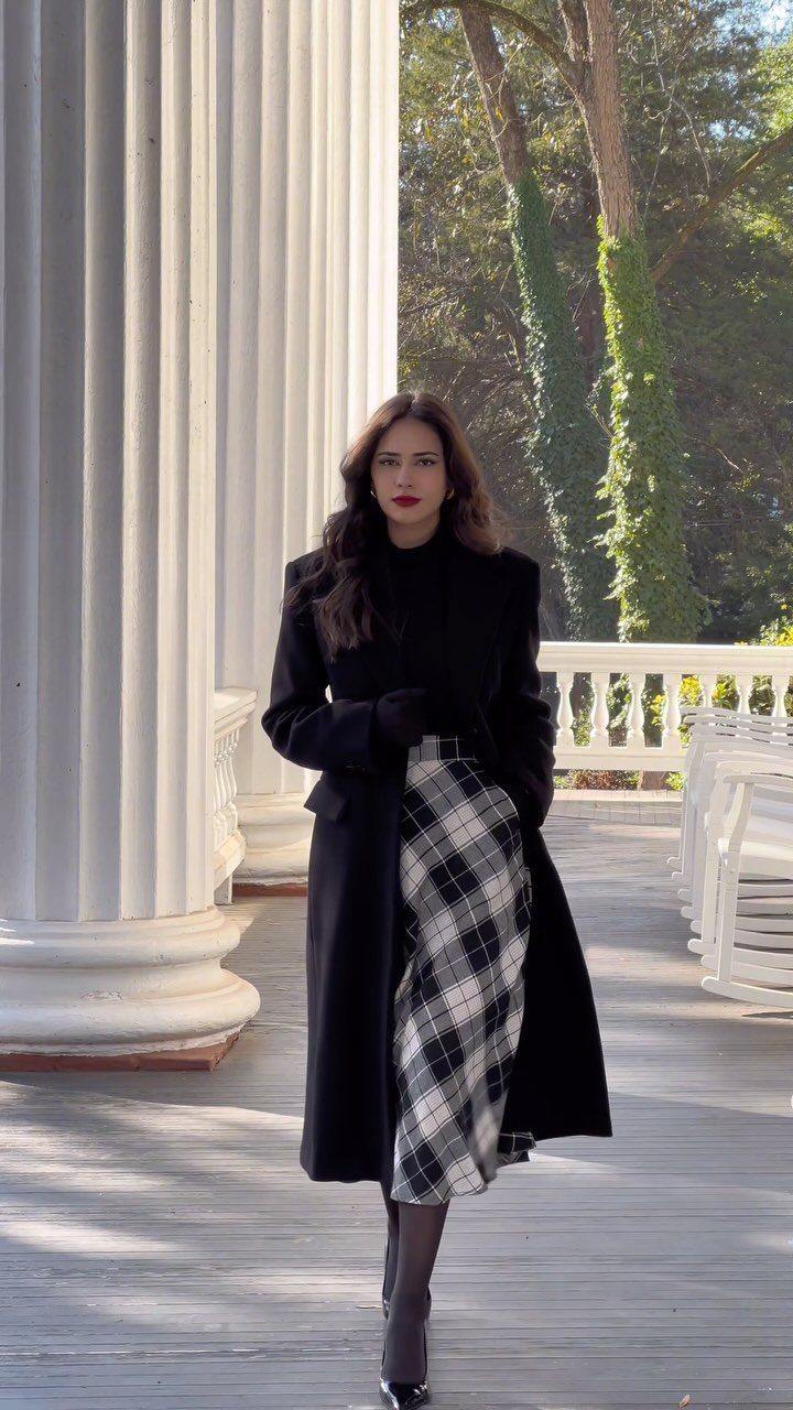 29 Fall Outfits Teens Can Copy, From Grunge to Cowgirl Looks 4 Woman wearing a black coat and plaid midi skirt standing by white columns.