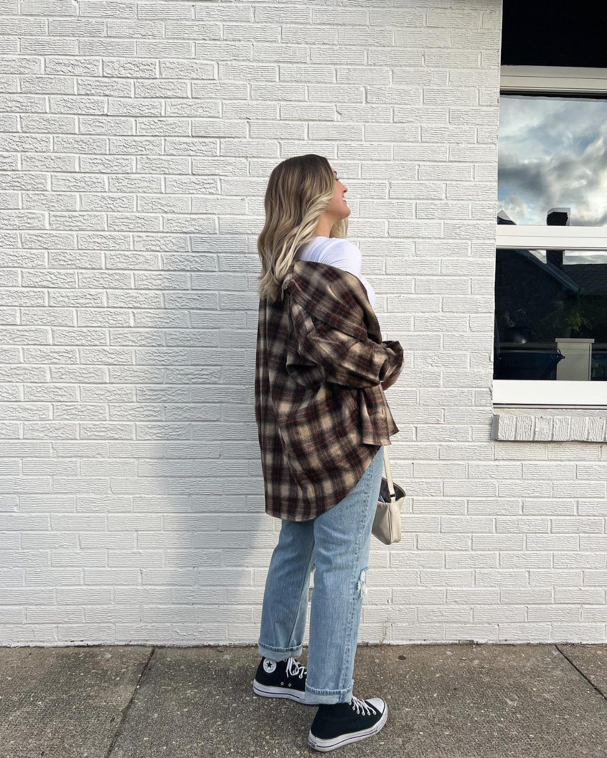 29 Fall Outfits Teens Can Copy, From Grunge to Cowgirl Looks 16 Person wearing an oversized brown plaid shirt and light blue jeans against white bricks.