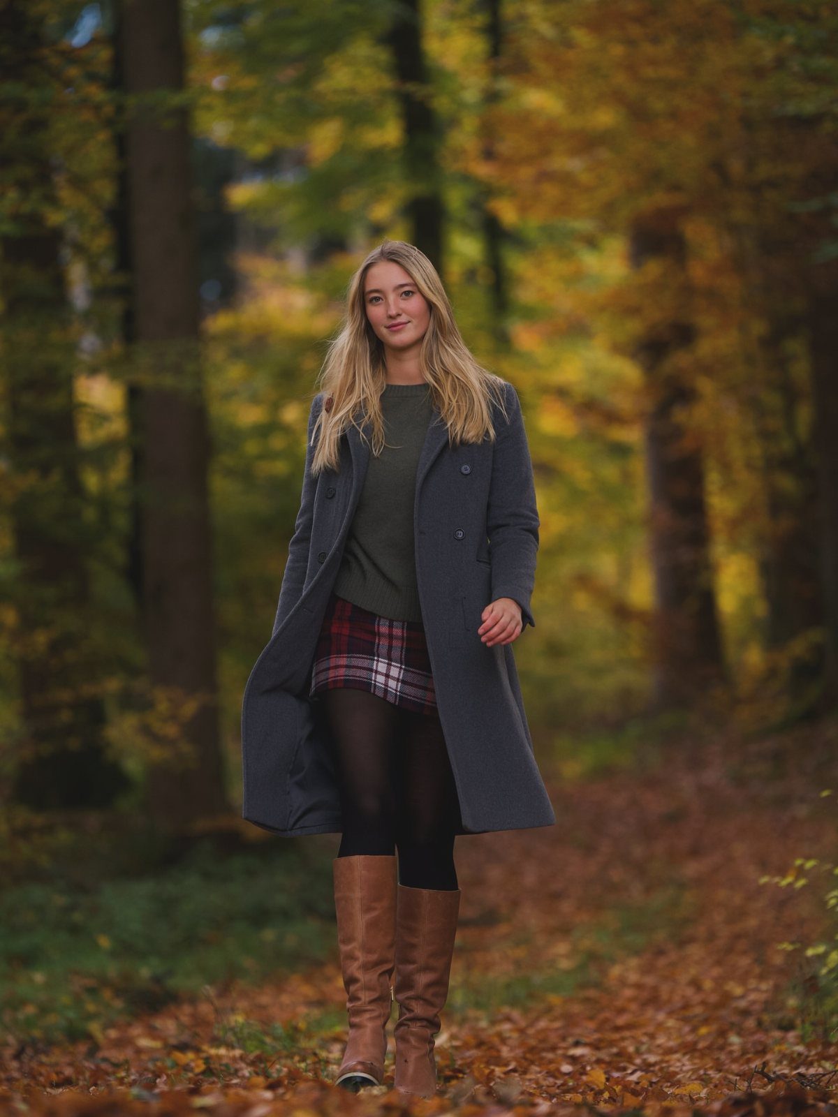 29 Fall Outfits Teens Can Copy, From Grunge to Cowgirl Looks 18 Young woman in a grey coat, green turtleneck, and plaid skirt in autumn forest.