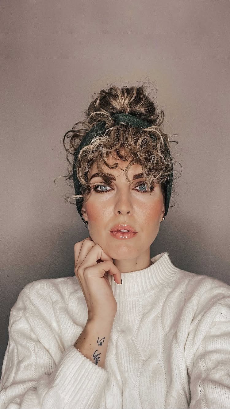 16 Good Hairstyles for Curly Hair, From Pixie to Long Layers 11 Woman with curly bangs and a bun wearing a white cable-knit sweater.