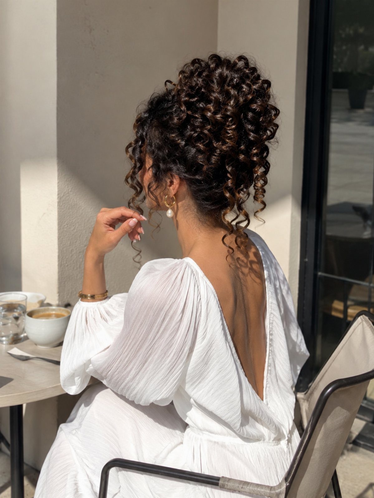 16 Good Hairstyles for Curly Hair, From Pixie to Long Layers 4 Woman with dark curly hair in a high messy bun wearing a white dress.