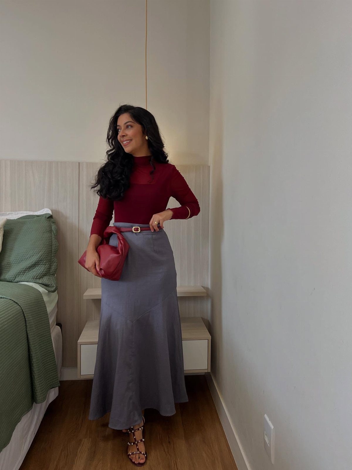 18 Gray Skirt Outfit Ideas From Mini to Maxi and Satin 12 Woman wearing a gray maxi skirt, burgundy top, and red handbag in a bedroom.