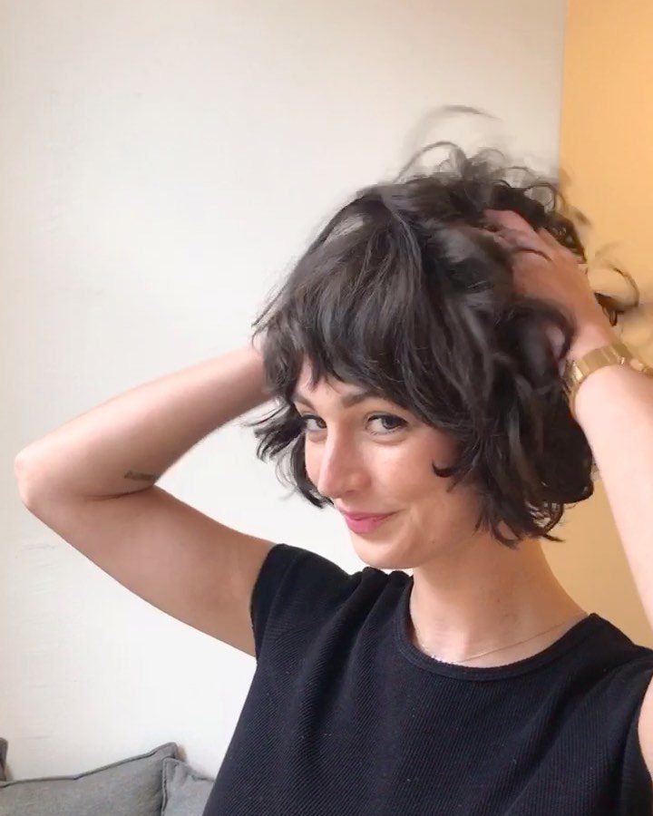 Woman with a dark curly bob and full bangs posing against a white wall