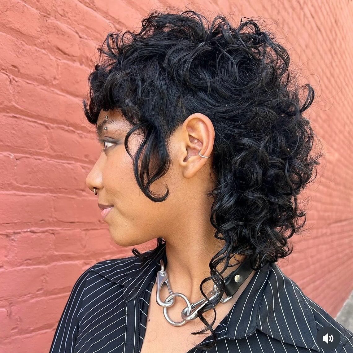 Person with a curly mullet haircut posing against a red brick wall.