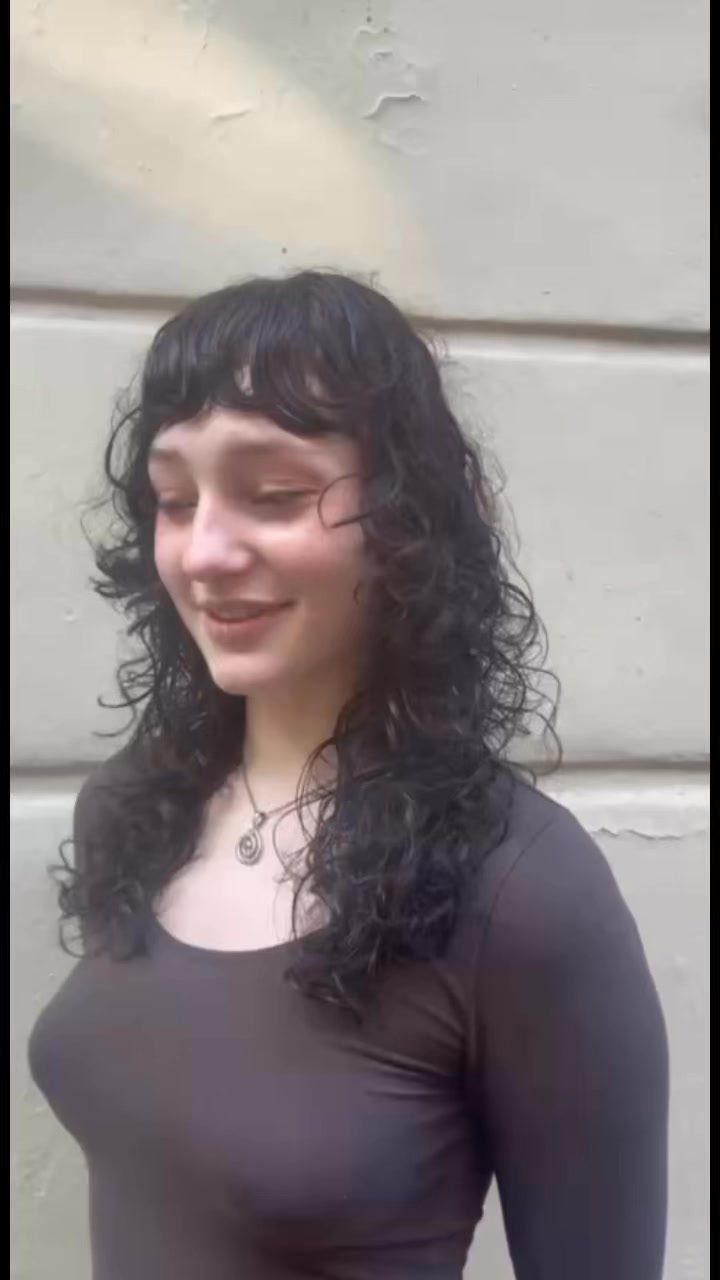 Woman with a curly shag haircut and bangs posing against a plain wall.