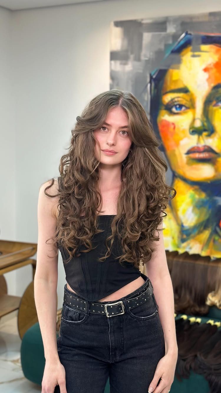 Woman with long brown curly hair and face-framing waves posing by a painting.