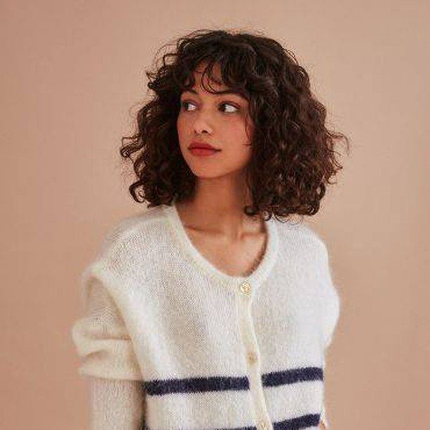 Woman with a curly layered lob and wispy bangs wearing a white cardigan.