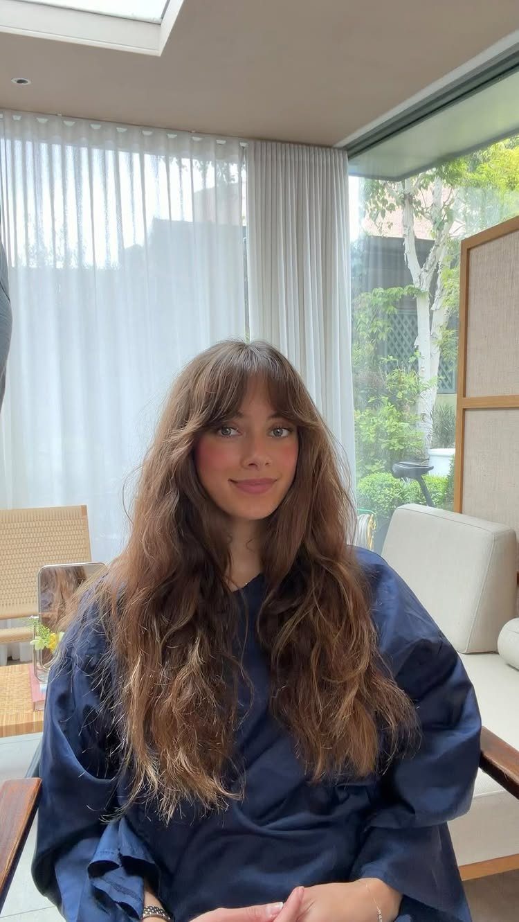 Woman with long wavy brown hair and soft curtain bangs posing indoors.