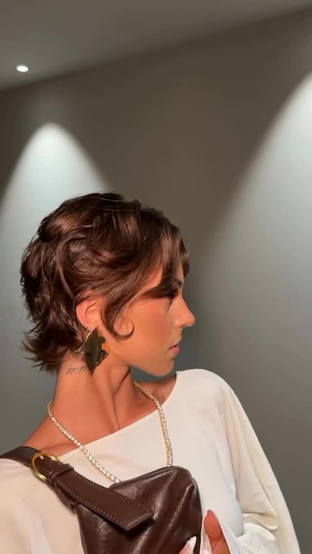 Woman with a short brown curly pixie bob haircut and gold earrings.