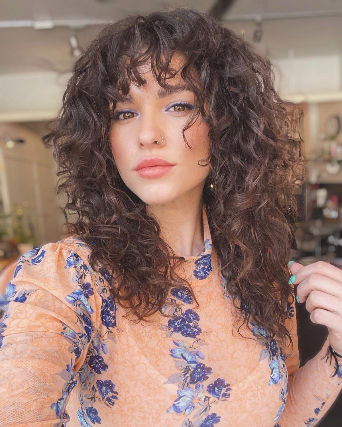 Woman with curly brown lob and wispy curly bangs wearing a floral blouse.