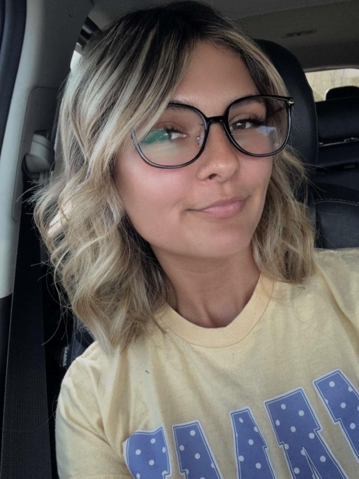 12 Hairstyles to Hide Bangs, From Quick Sweeps to Sleek Buns 6 Woman with wavy blonde highlighted hair and middle-parted face-framing bangs wearing glasses.