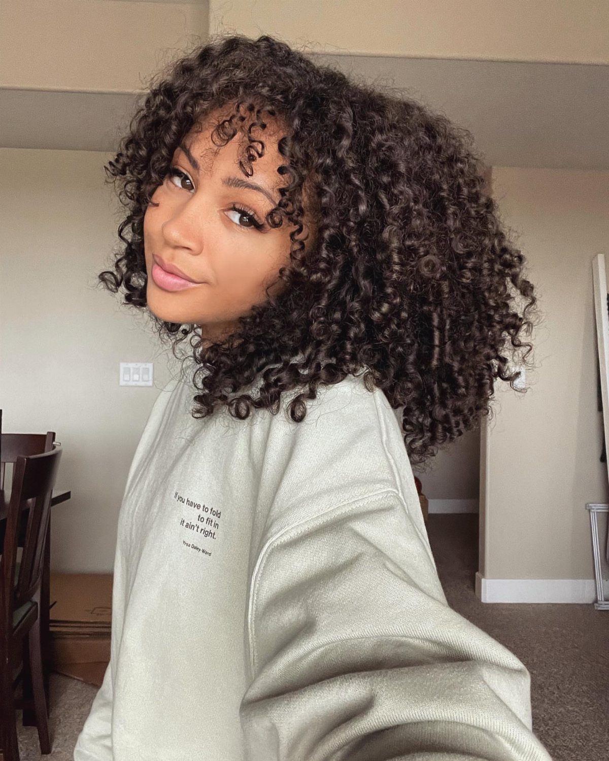 Woman with voluminous curly black hair and bangs wearing a grey sweatshirt.