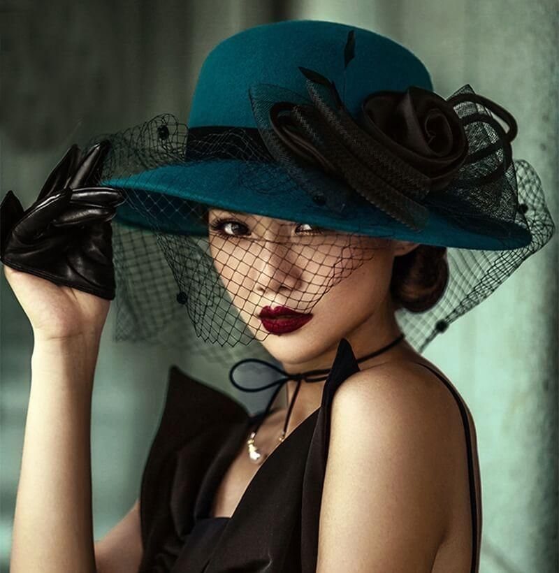 Woman wearing a teal wide-brimmed hat with a black rose and net veil.