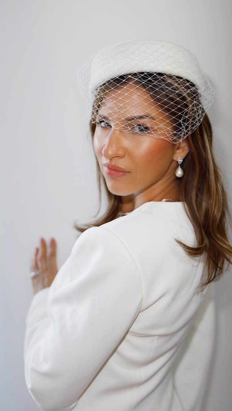 Woman wearing a white pillbox hat with a net birdcage veil and white blazer.