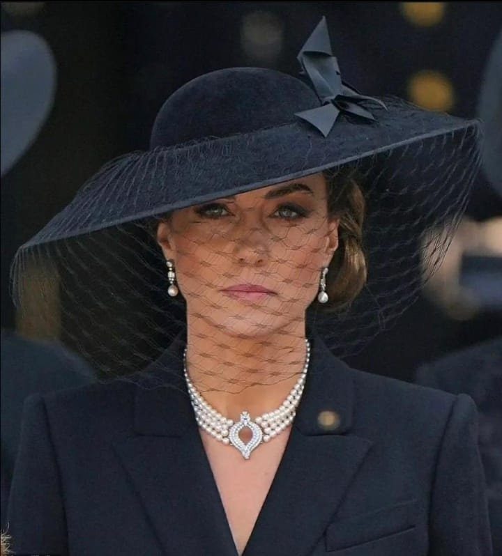 Woman wearing a wide-brimmed black hat with a net veil and pearl jewelry.